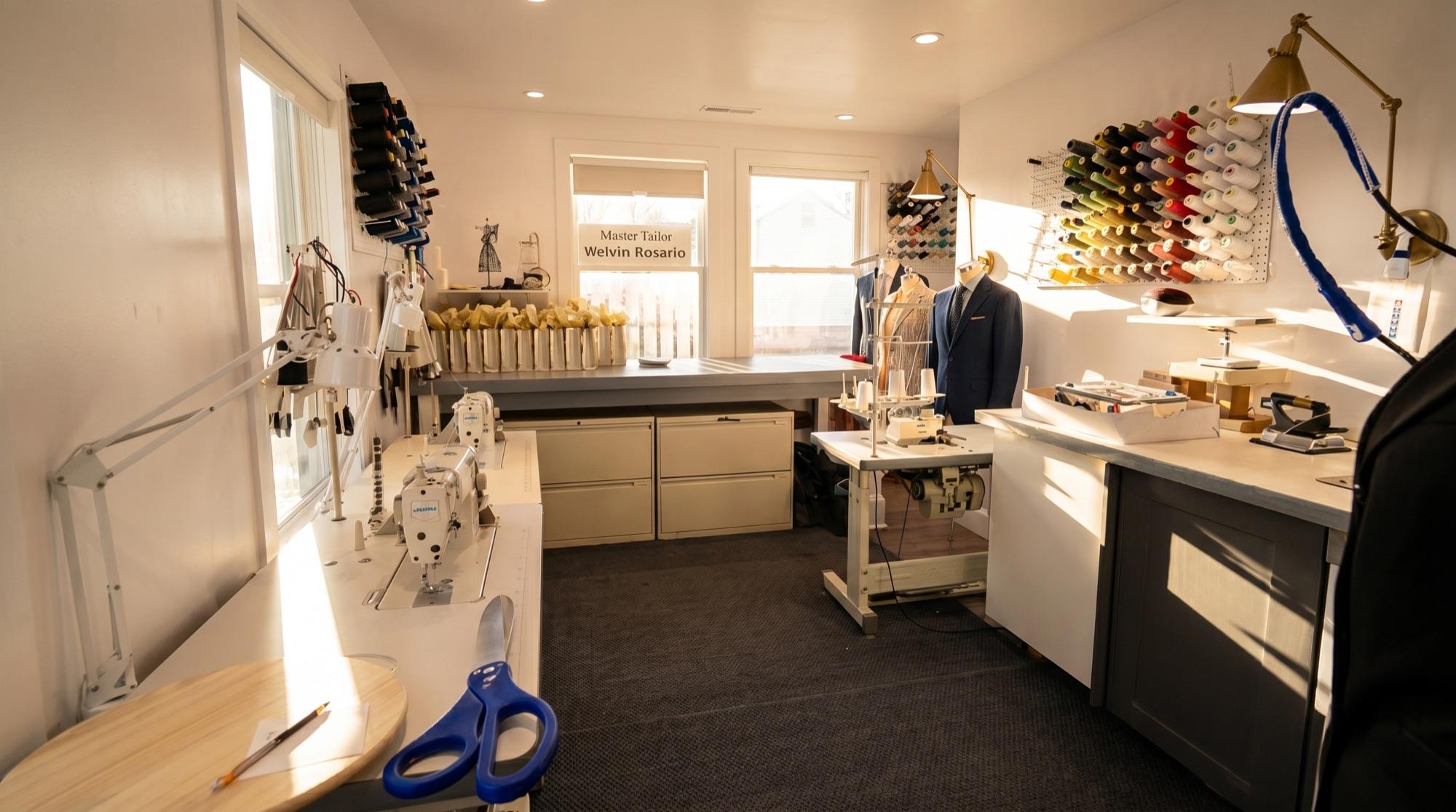 Wide view of the Westport atelier interior — cutting table, sewing machines, thread rack, and Welvin Rosario Master Tailor sign in natural window light