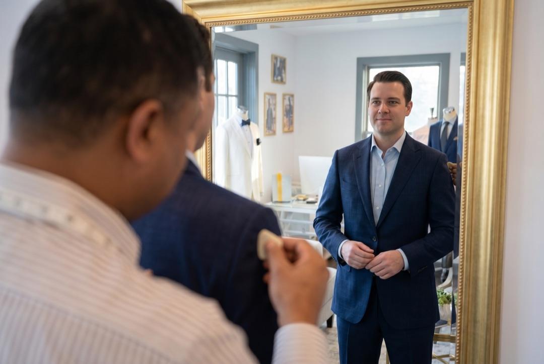 Client buttoning his finished navy suit jacket in the gilt-framed fitting mirror, seen over Welvin's shoulder