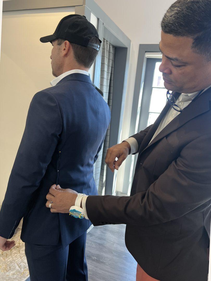 Welvin pinning the back of a navy jacket on a standing client, pincushion strapped to his wrist, atelier window light behind them