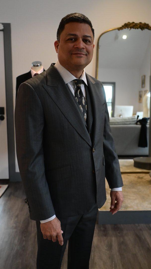 Client wearing a charcoal three-piece bespoke suit with peak lapels, matching vest, white dress shirt, and patterned silk tie, atelier mirror and dress form behind